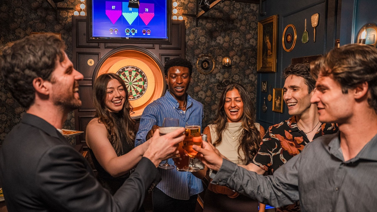 Group of friends toasting with drinks near a dartboard in a lively bar setting.