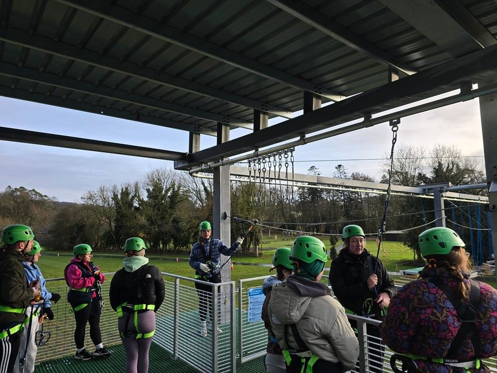Group of people in harnesses and helmets at a zipline platform outdoors.