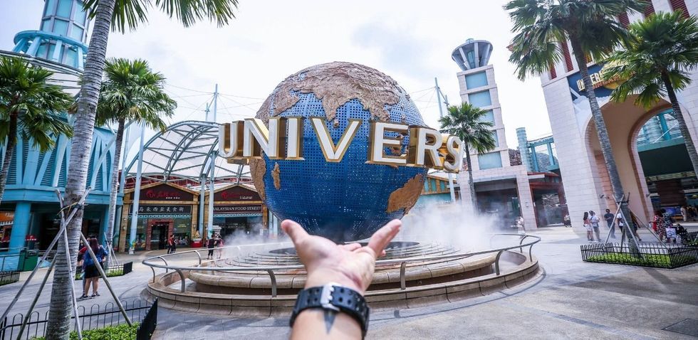 Hand gestures towards Universal Studios globe amid palm trees and buildings.