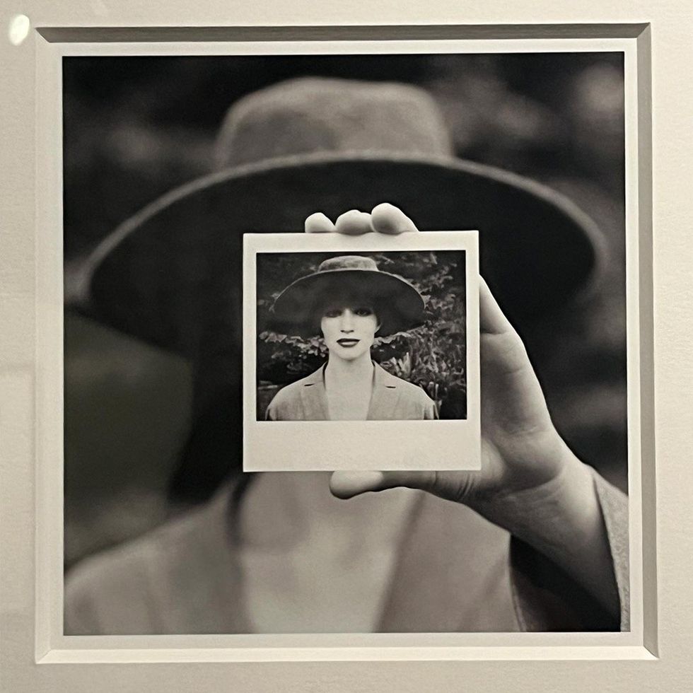 Hand holding a Polaroid of a person wearing a hat, matching the background figure.