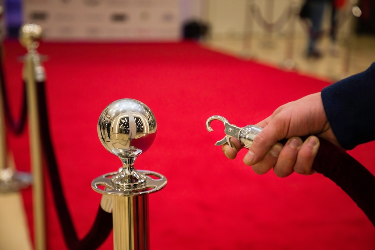 Hand holding a stanchion hook by a red carpet on a polished metal post.