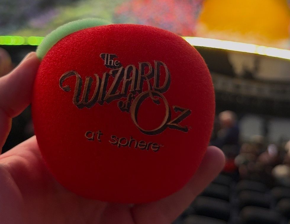 Hand holding red "Wizard of Oz at Sphere" themed item, blurred background.