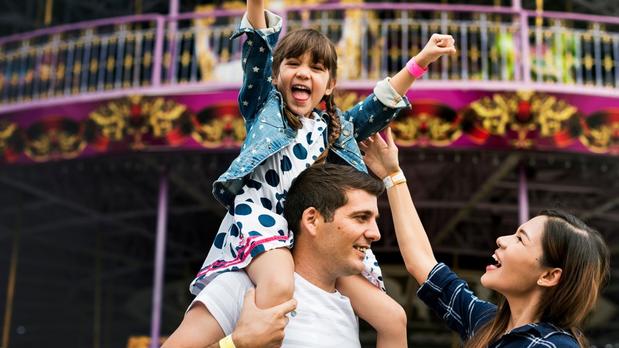 Happy family at a colorful amusement park, child on dad's shoulders, mom cheering.