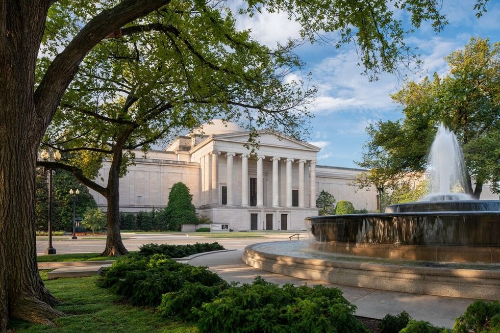 Historic building called the National Gallery of Art with columns, surrounded by trees, near a fountain under a blue sky.