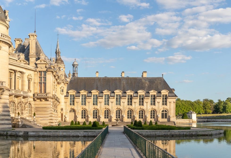 Historic castle, Ch\u00e2teau de Chantilly, with ornate architecture and a reflecting pond under a blue sky.