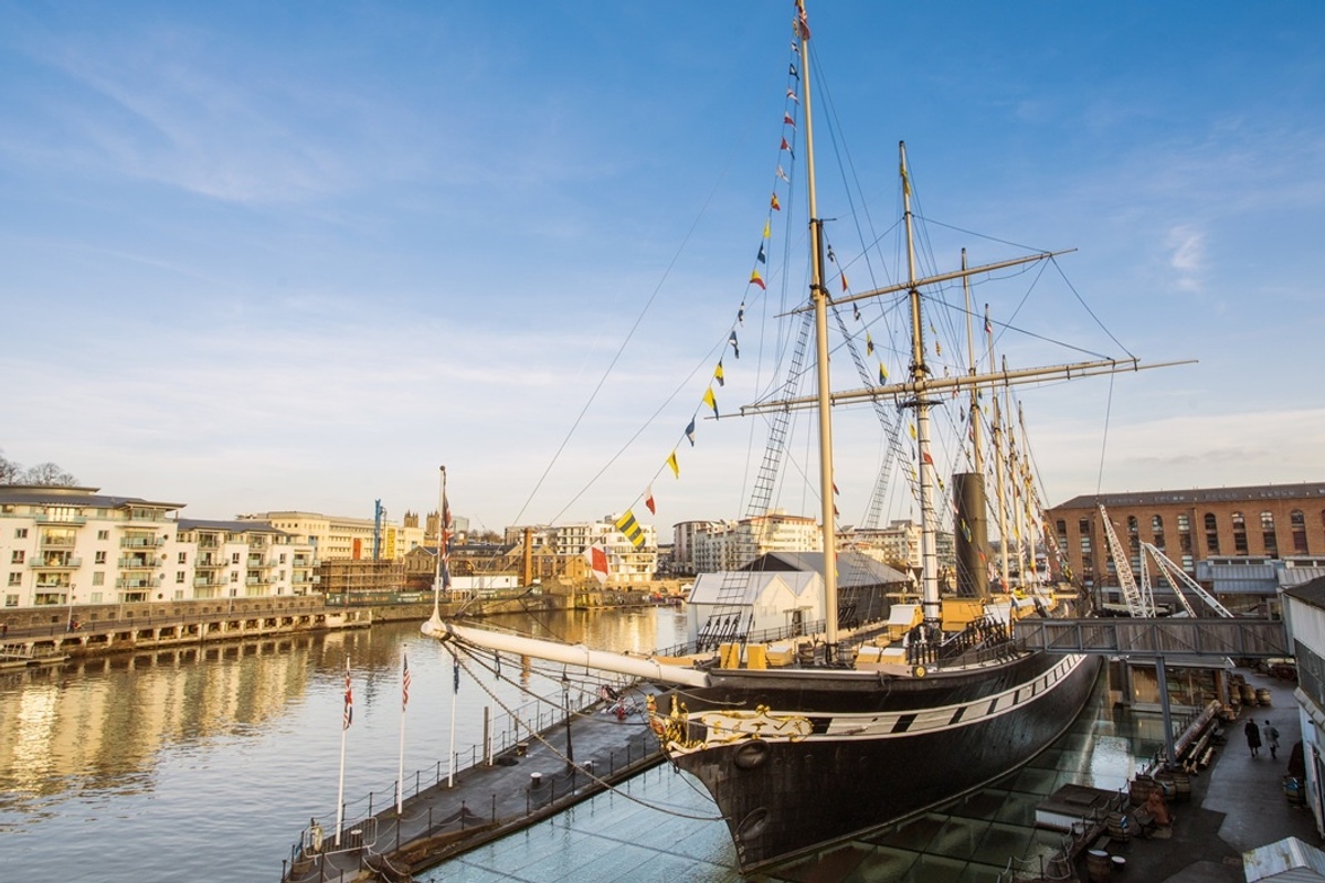 Historic sailing ship at Bristol Dockyards, by waterfront buildings under a clear blue sky.