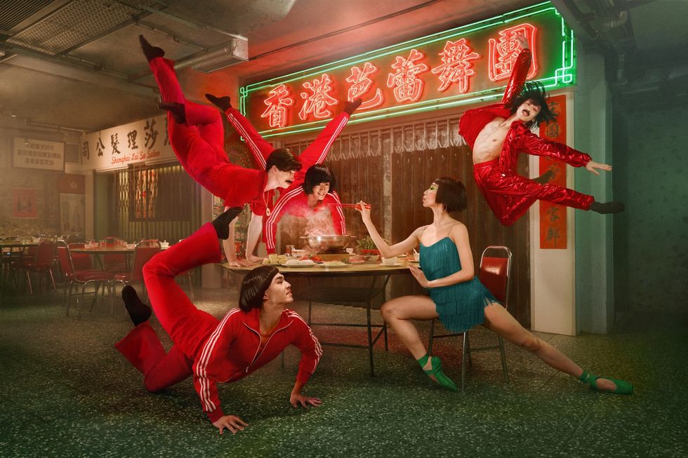 Hong Kong Ballet dancers in red jumpsuits perform energetically around a dining table in a neon-lit restaurant.