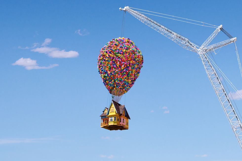 House lifted by colorful balloons attached to a crane under a clear blue sky.