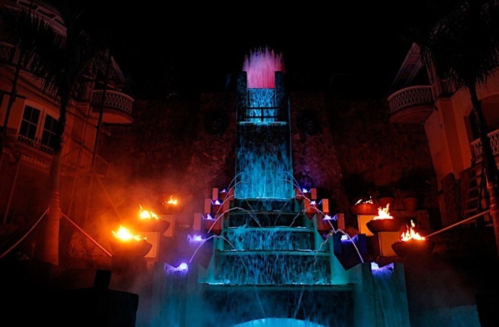 Illuminated waterfall at night with fire bowls and vibrant blue and orange lights.