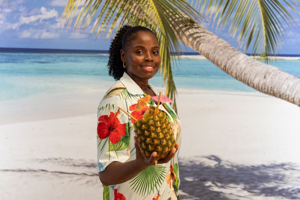 Immersive decor at Suntago Water World - a woman holding pineapple drink, standing on a beach with palm trees in the background.