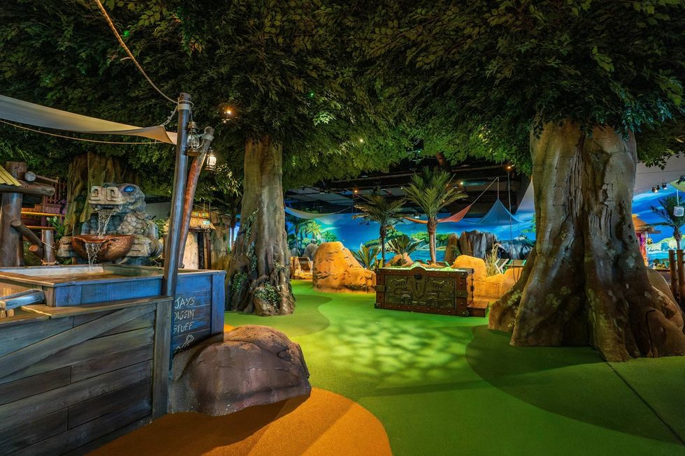 Indoor mini-golf course with large artificial trees and a treasure chest in a jungle theme.