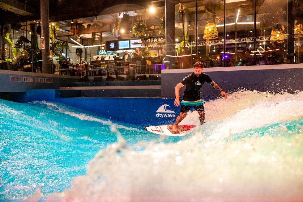 Indoor surfer rides artificial wave; onlookers sit in bar above.