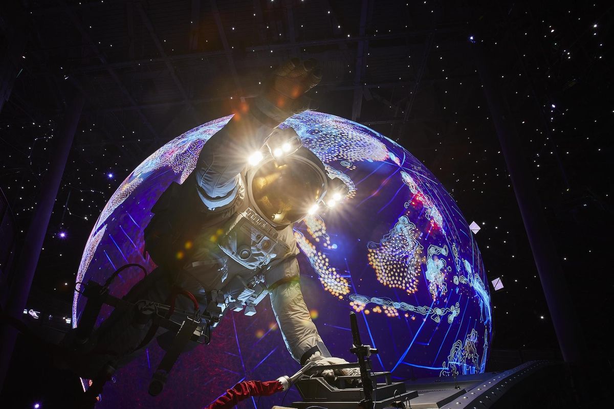 Inside Alif – The Mobility Pavilion, Astronaut reaches towards a colorful, illuminated Earth model in a starry, dark setting.