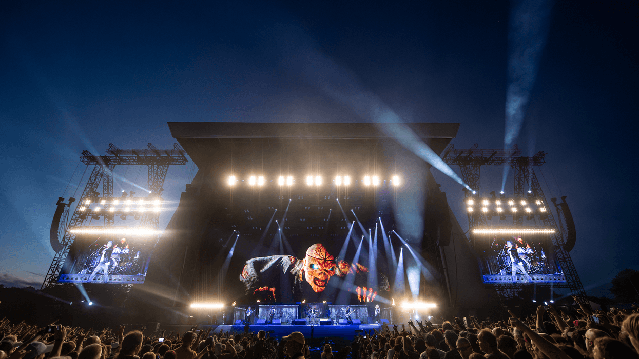 Iron Maiden concert stage with giant screens, lights, and a large crowd at dusk.