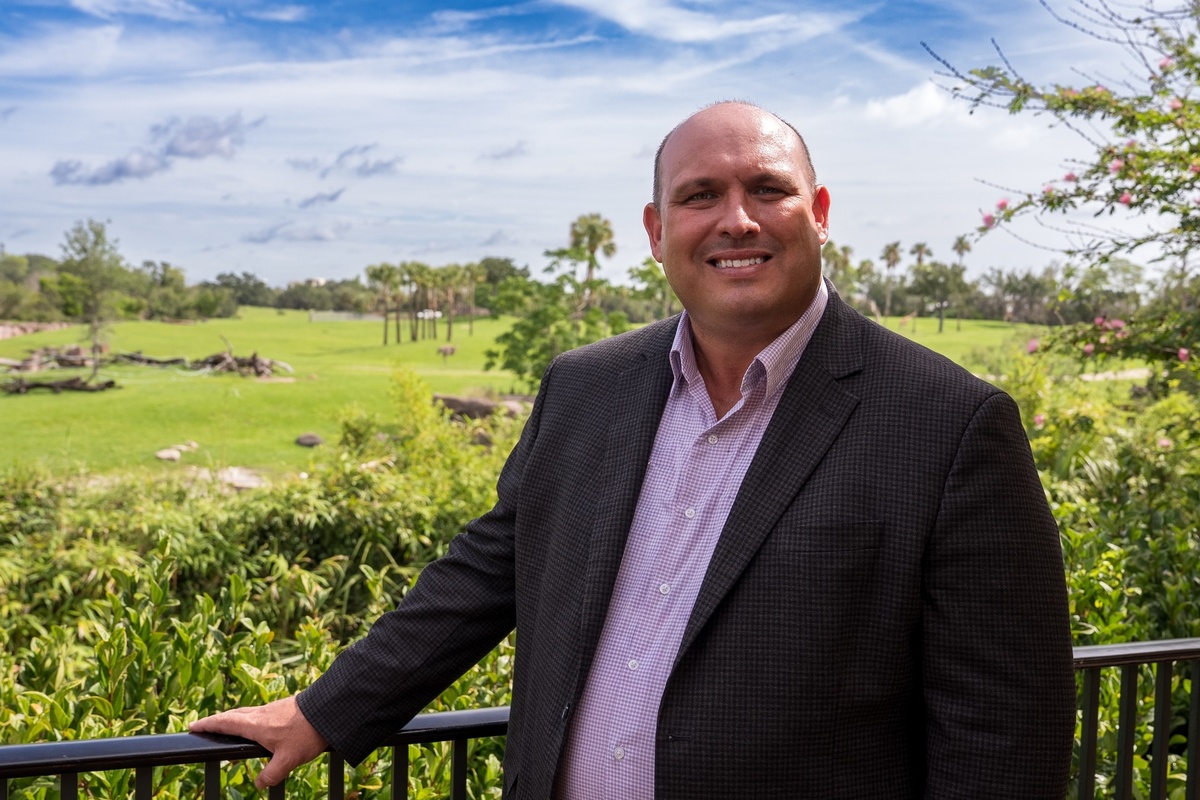 Jon Vigue in a suit smiling outdoors with a scenic green landscape in the background at Busch Gardens Tampa Bay