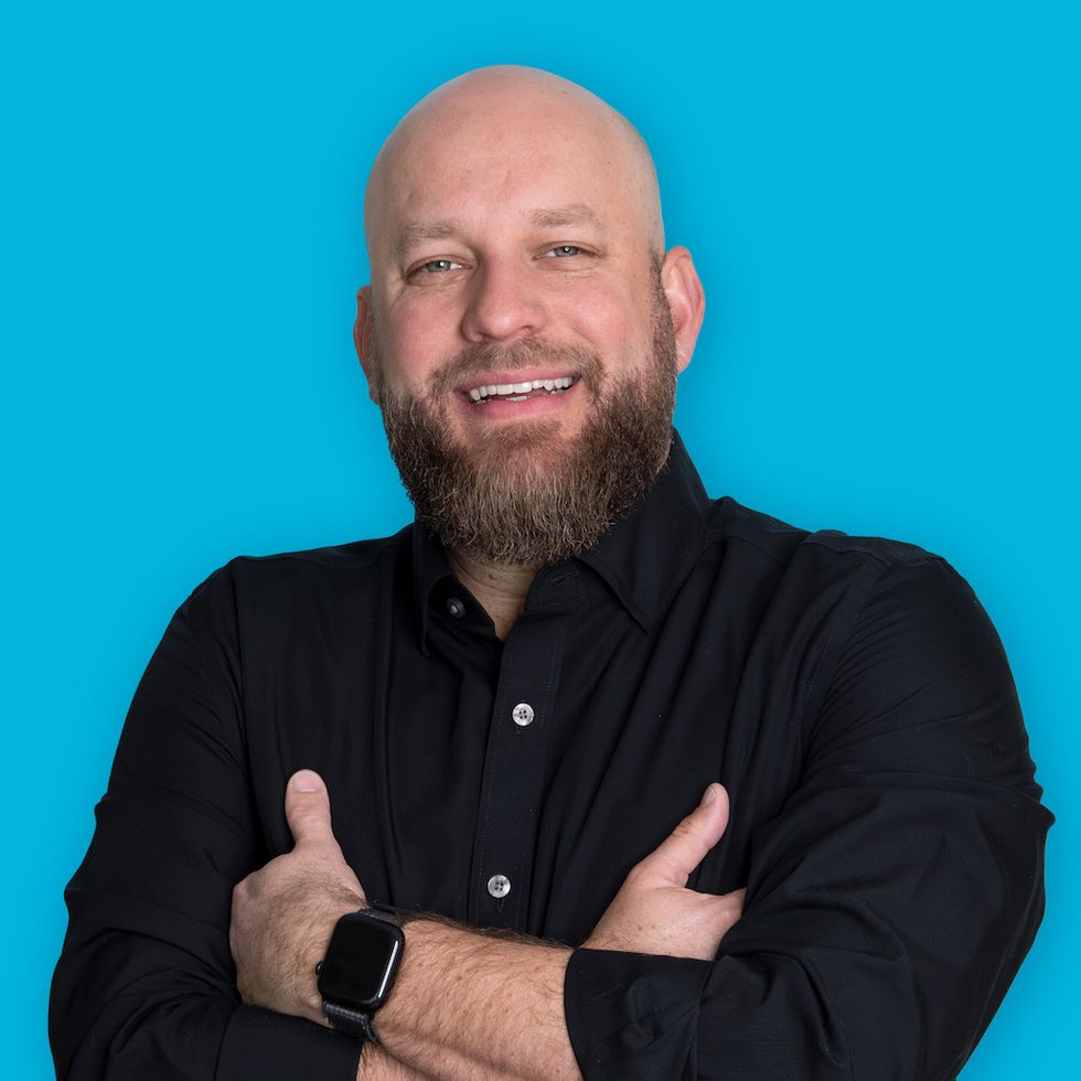 Josh Martin, Smiling man with a beard, crossing arms, wearing a black shirt, blue background.