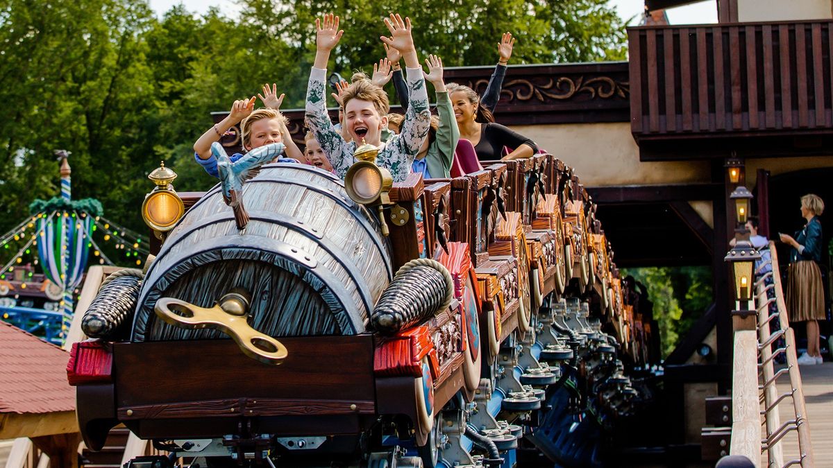 Kids enjoying a roller coaster ride with hands raised, surrounded by trees.