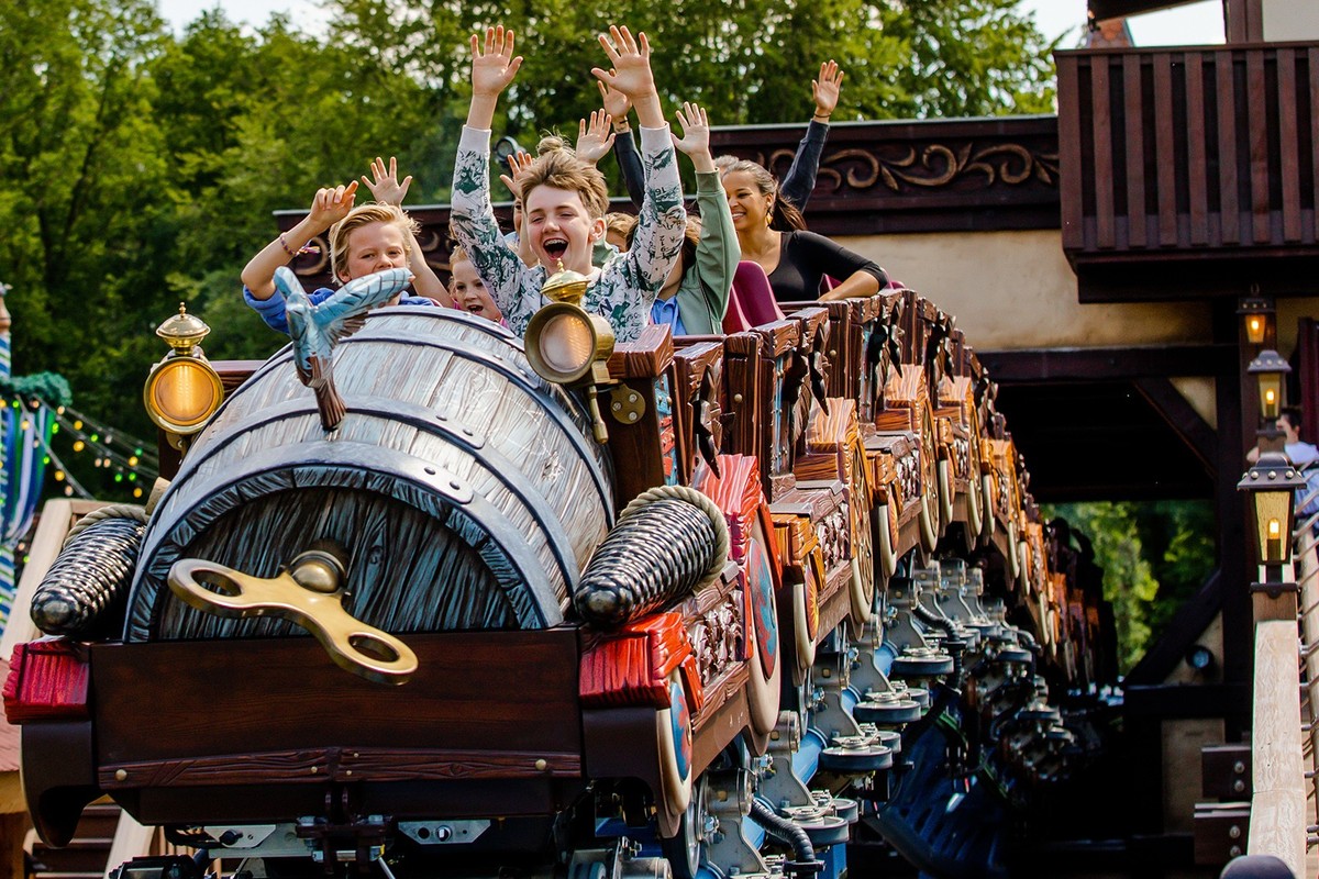Kids enjoying a roller coaster ride with hands raised, surrounded by trees.