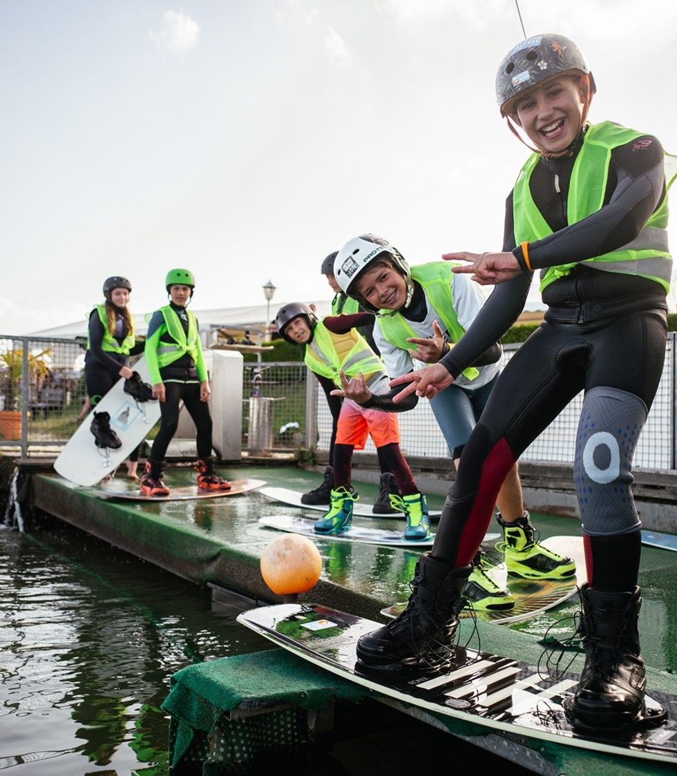 Kids in helmets and vests ready for wakeboarding, smiling and posing on a platform by the water.