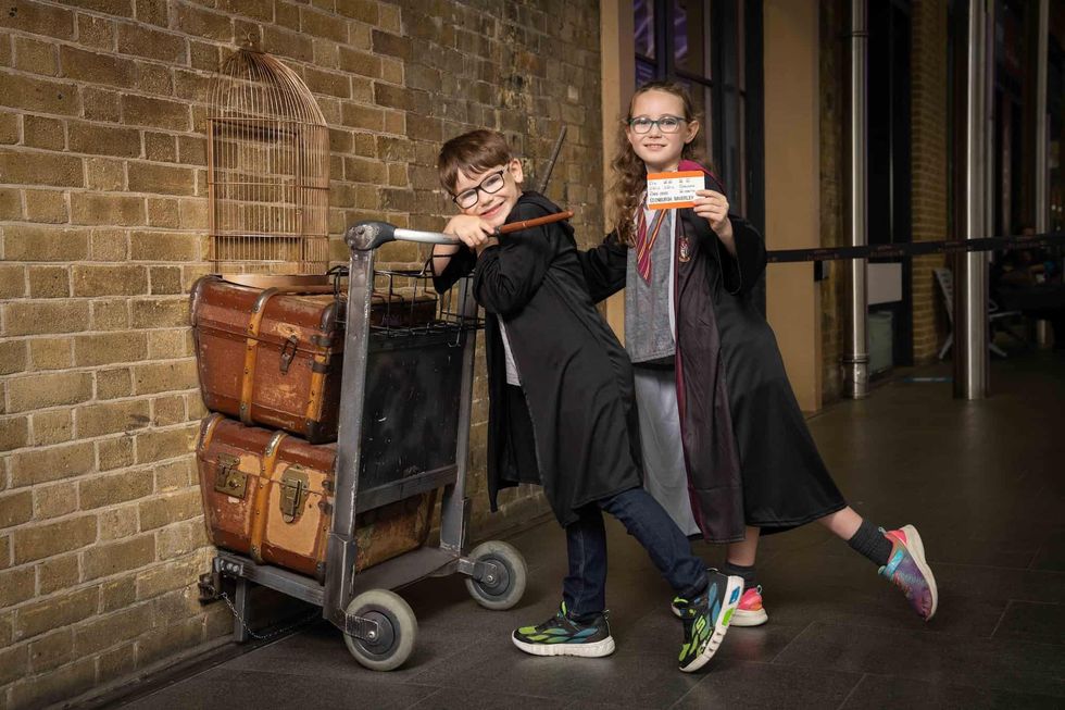Kids in wizard robes pose with luggage cart at a brick wall, holding a train ticket.