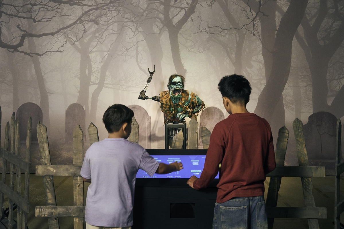 Kids interact with a touchscreen, facing a spooky animatronic in a graveyard setting at the Academy Museum
