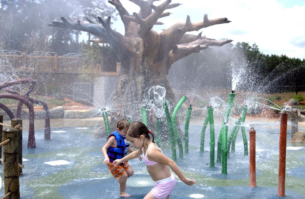 Kids playing at a splash pad with water features and a large tree in the background.