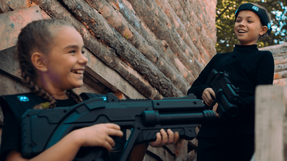 Kids playing laser tag, smiling and holding toy guns, against a wooden background.