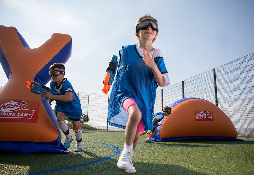 Kids playing Nerf battle at outdoor training camp with inflatable obstacles.