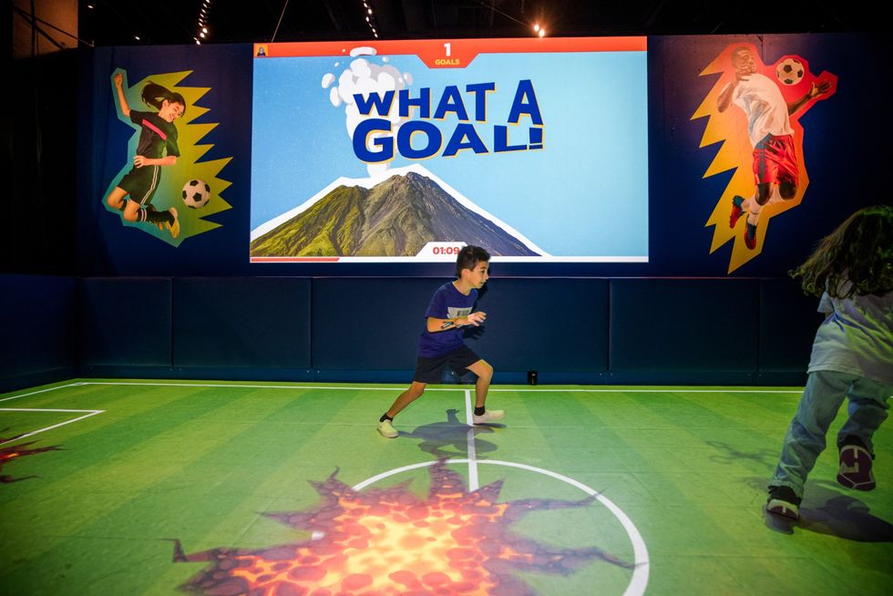Kids playing soccer in an interactive exhibit with a "What a Goal!" screen.