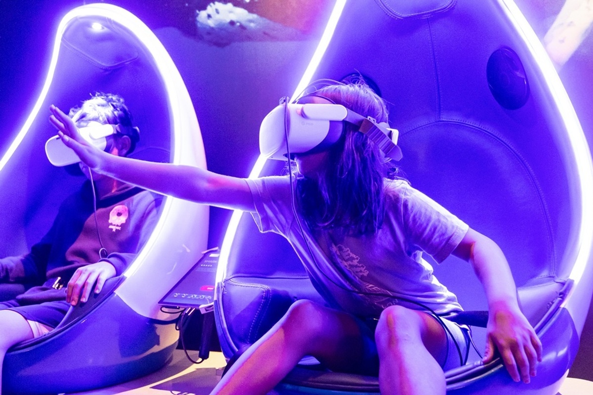Kids wearing VR headsets in futuristic gaming chairs in an Immotion experience, reaching out towards a virtual scene.