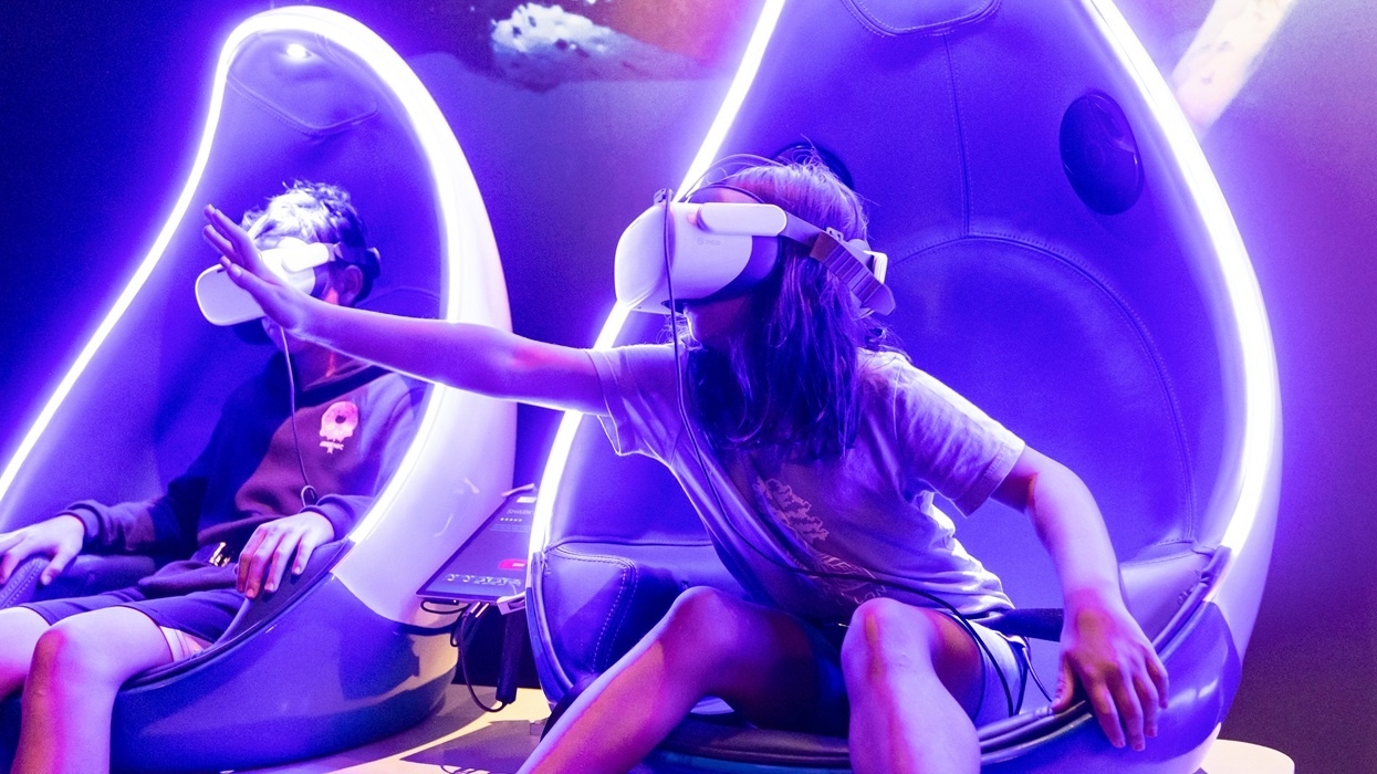 Kids wearing VR headsets in futuristic gaming chairs in an Immotion experience, reaching out towards a virtual scene.