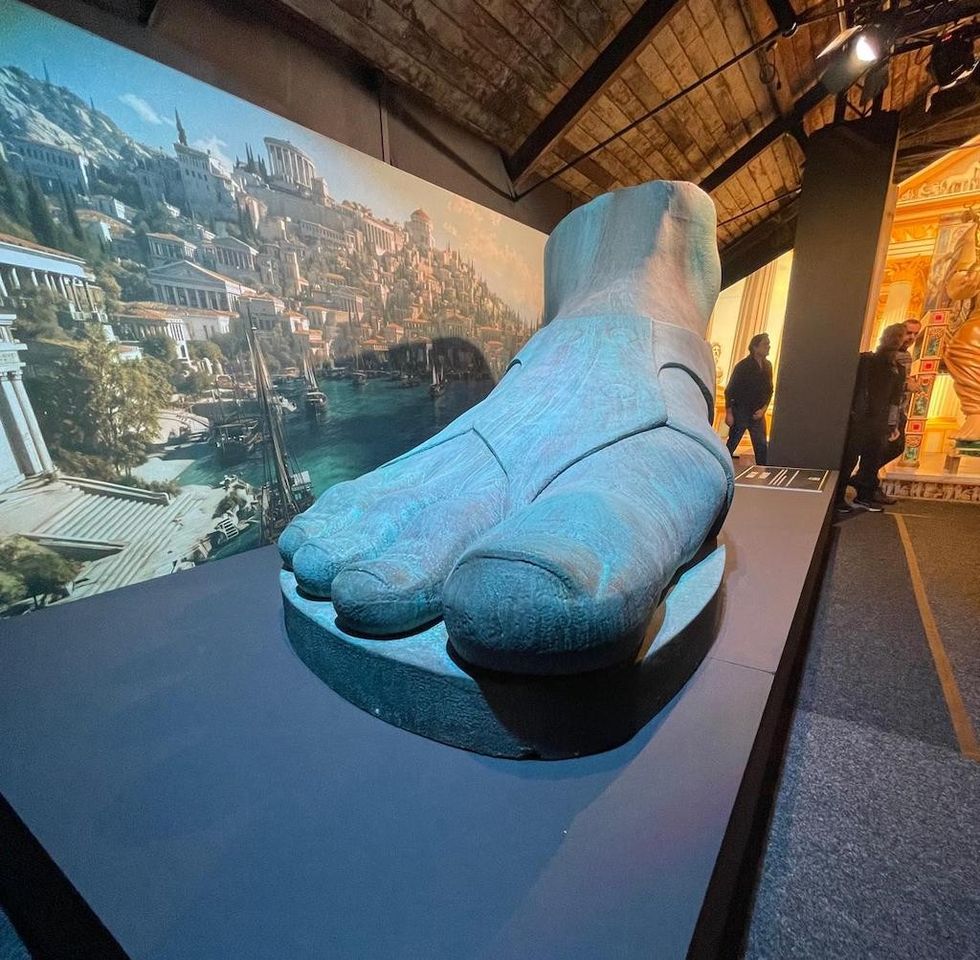 Large ancient statue foot with detailed backdrop of classical cityscape.