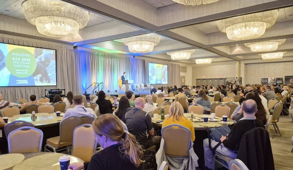 Large conference with a speaker on stage and attendees seated at round tables.