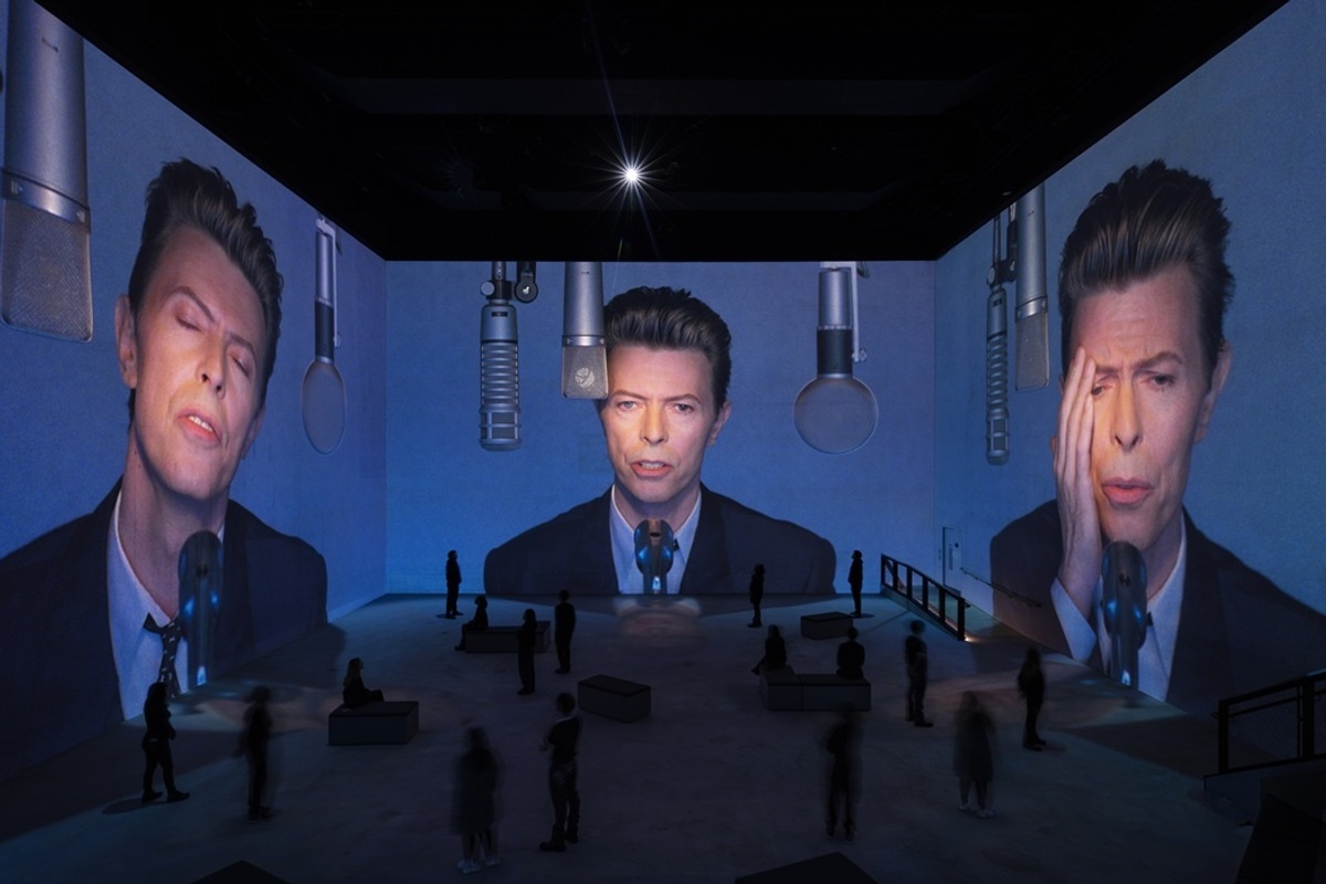 Large projections of David Bowie's face on walls, with people observing in a dimly lit room at Lightroom, London