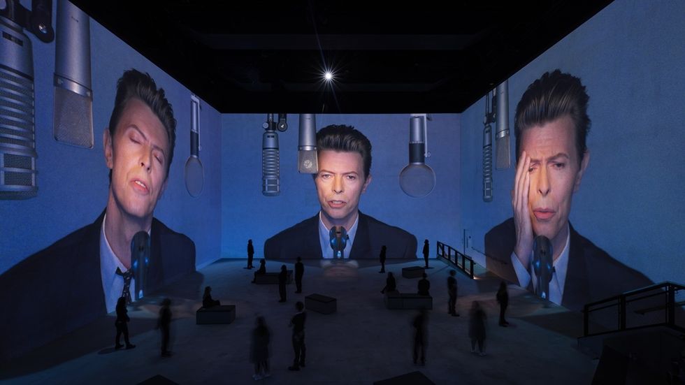 Large projections of David Bowie's face on walls, with people observing in a dimly lit room at Lightroom, London