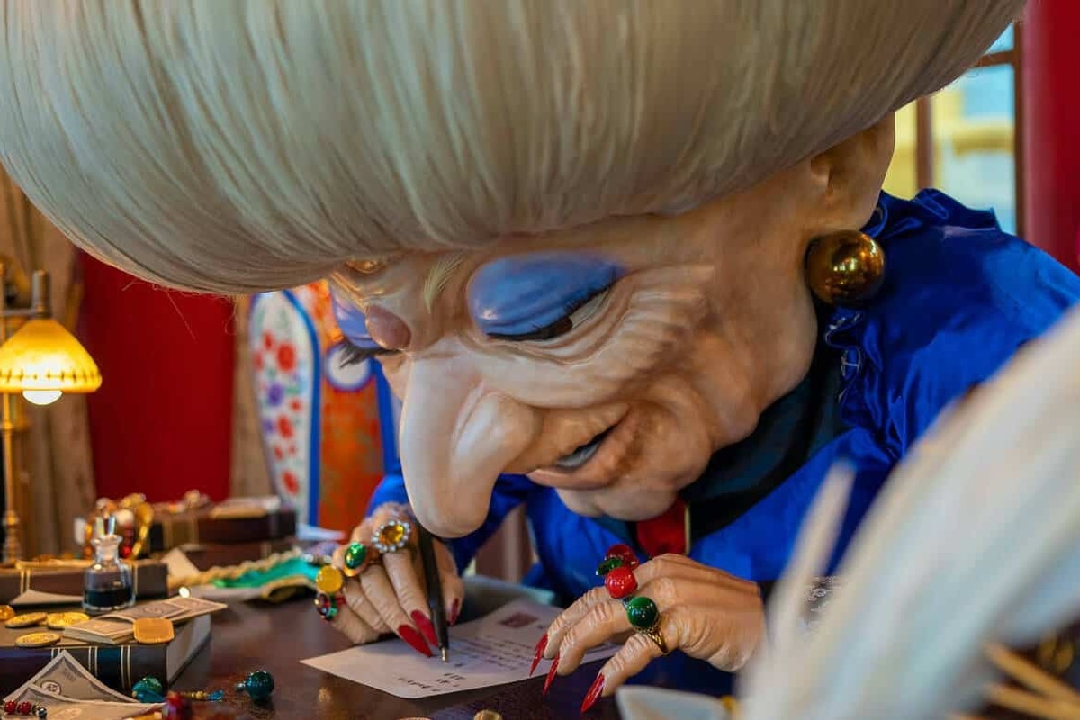 Large Studio Ghibli puppet, Yubaba from the film Spirited Away, with elaborate hair writes at a desk, wearing colourful rings and blue garments.