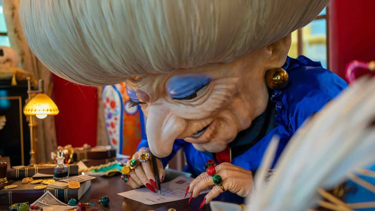 Large Studio Ghibli puppet, Yubaba from the film Spirited Away, with elaborate hair writes at a desk, wearing colourful rings and blue garments.