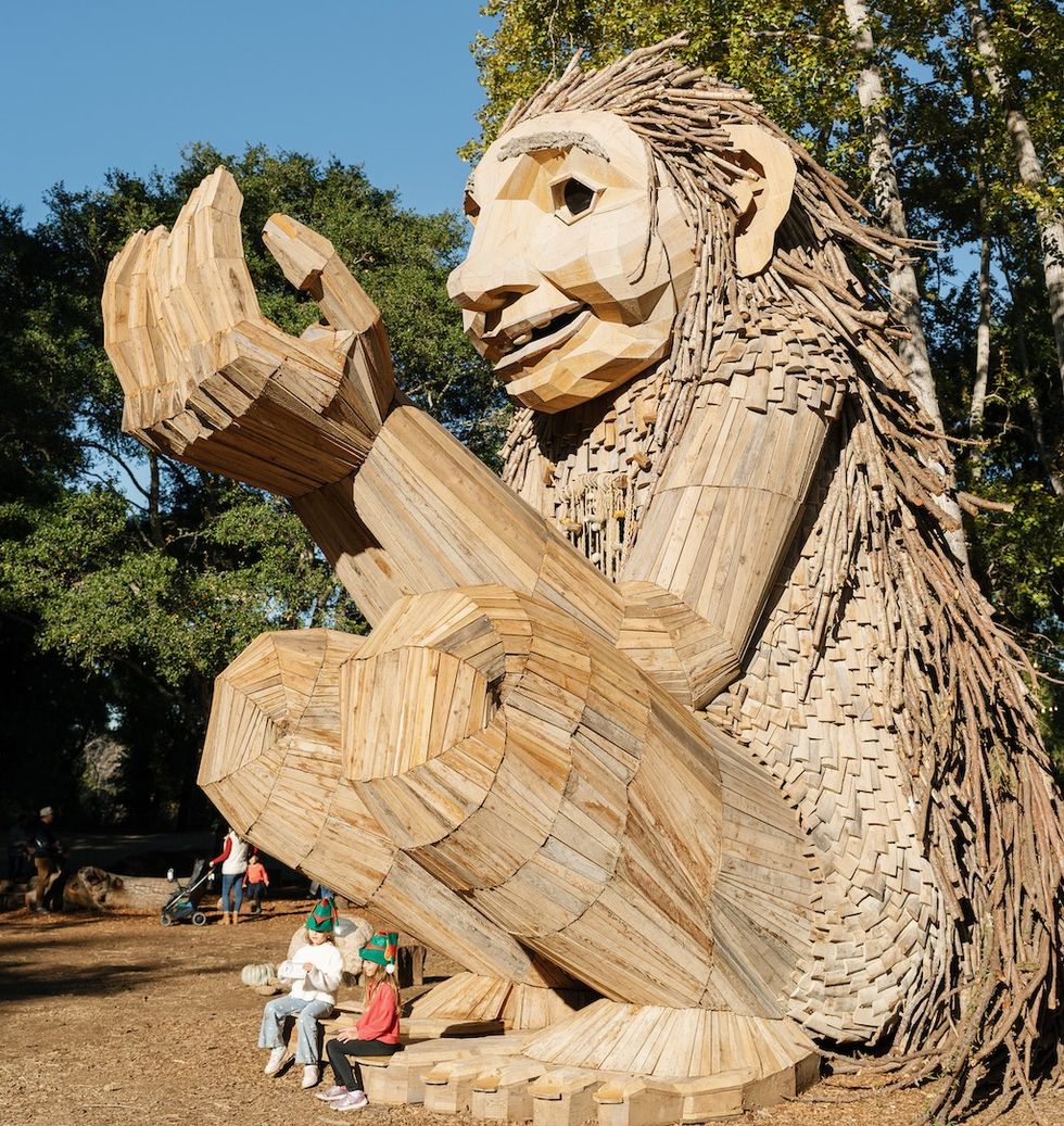 Large wooden troll sculpture holding two children on its arm in a forest setting.