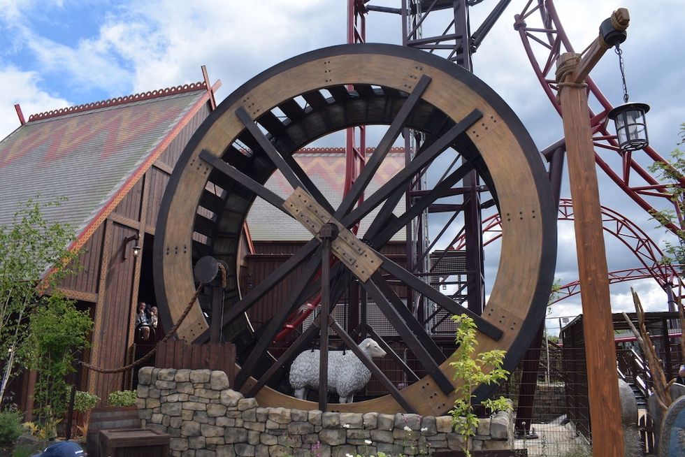 Large wooden waterwheel beside rustic buildings and roller coaster track, with sheep inside the wheel, Drakon coaster Paultons Park