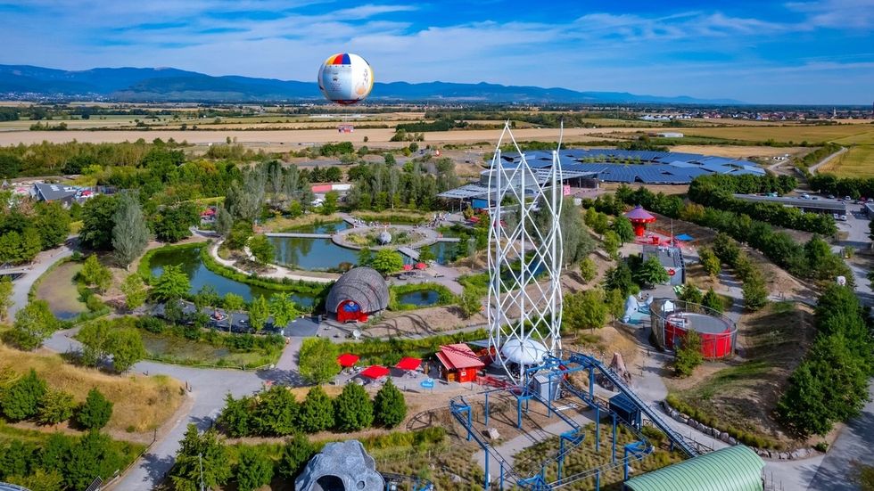 Le Parc du Petit Prince aerial view with tall ride, greenery, and distant hot air balloon.