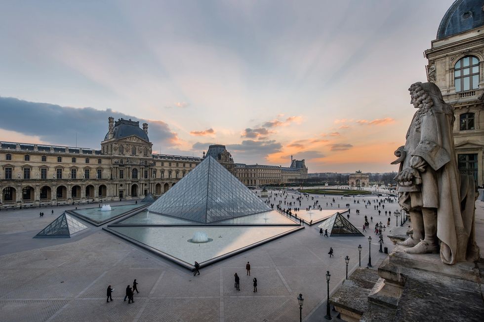 Louvre Pyramid at sunset, with people walking and a statue overlooking the scene.