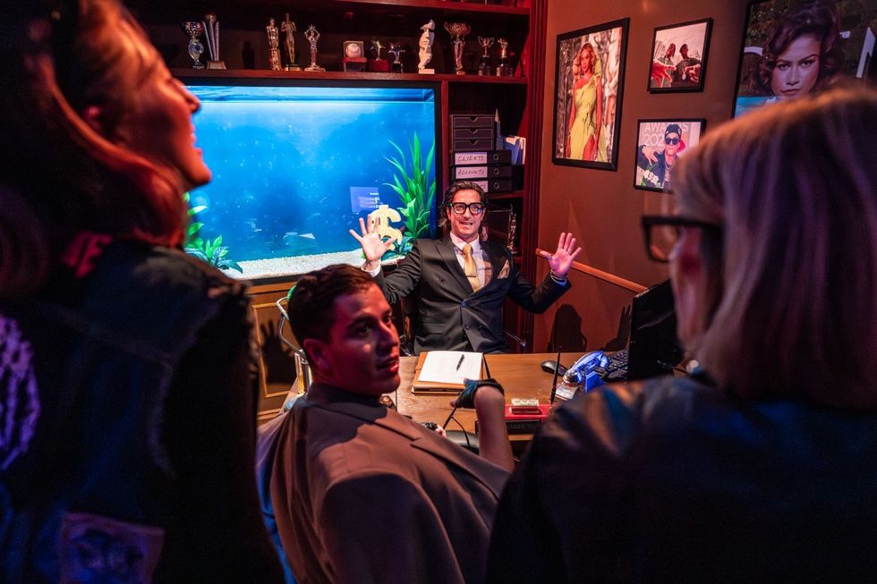 Man at a desk gesturing, surrounded by people and aquarium backdrop.