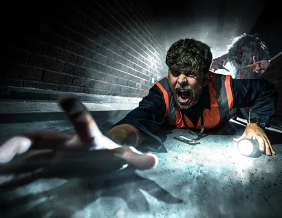 Man crawling on the ground, reaching for something, with a flashlight and zombie in the background.