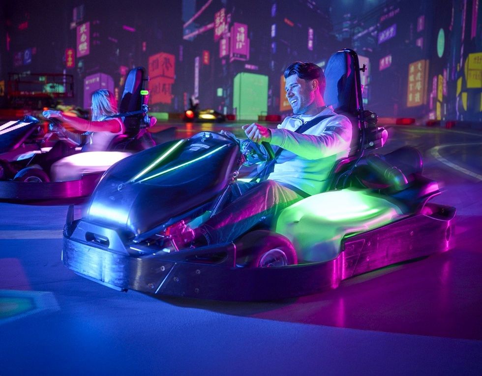 Man driving a neon-lit go-kart in an indoor arena with colorful, futuristic city backdrop.
