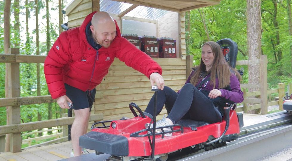 Man helping woman in a red sled at a forest ride station, both smiling.
