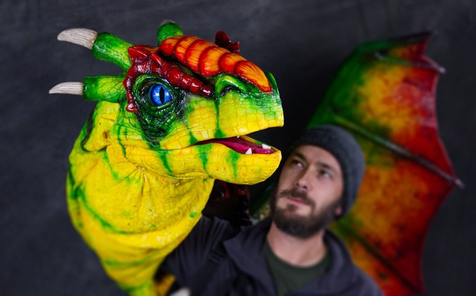 Man holds a colorful dragon puppet with green and red scales and blue eyes.
