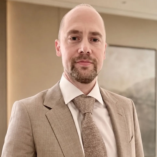 Man in a beige suit and tie, standing in a softly lit room.