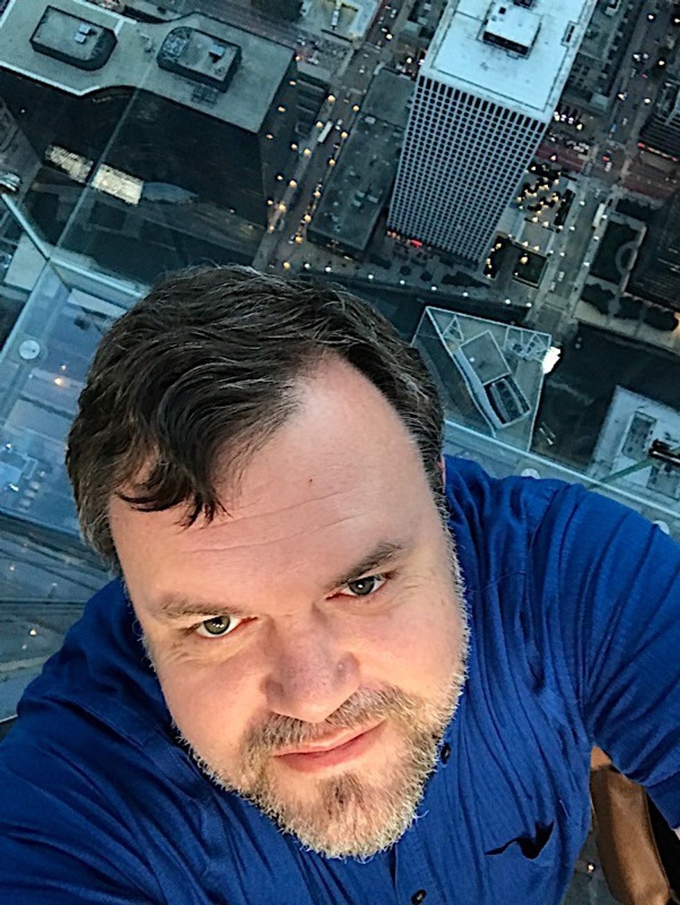 Man in a blue shirt taking a selfie high above a cityscape.