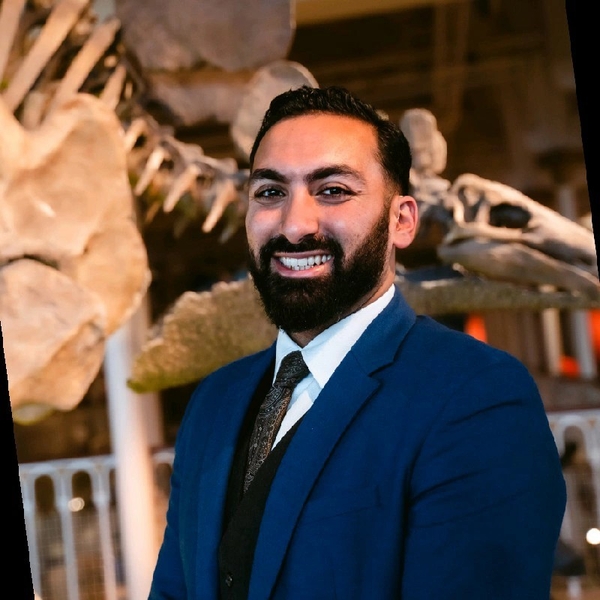 Man in a blue suit smiling in front of dinosaur skeleton display.