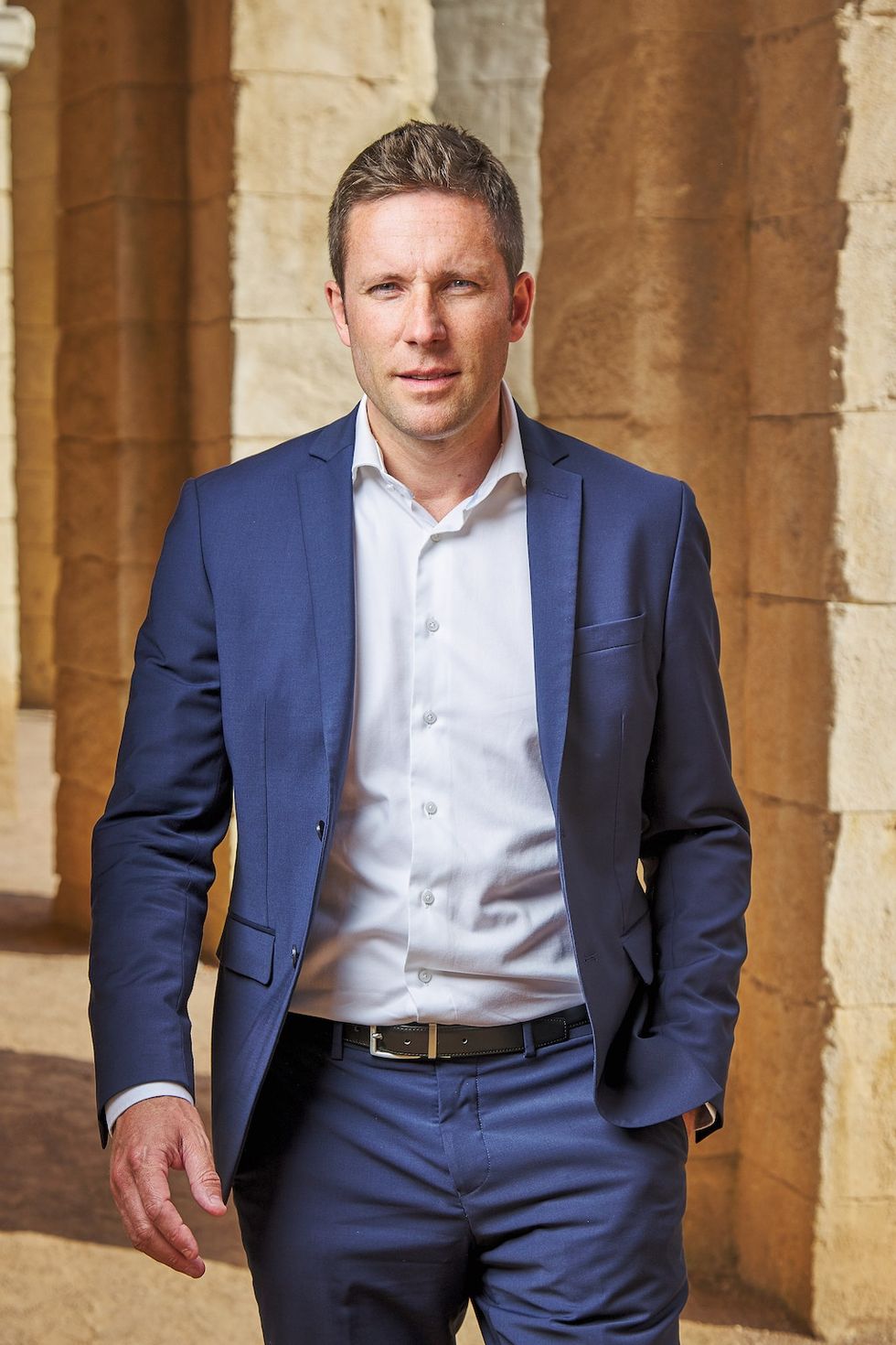 Man in a blue suit, \u200bNicolas de Villiers, walking through a stone archway.
